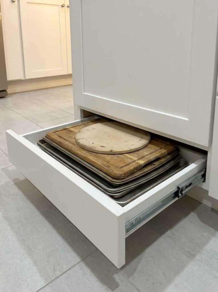 A close-up shot of a kitchen base cabinet at floor level, a toe-kick drawer pulled fully open revealing flat baking sheets,
